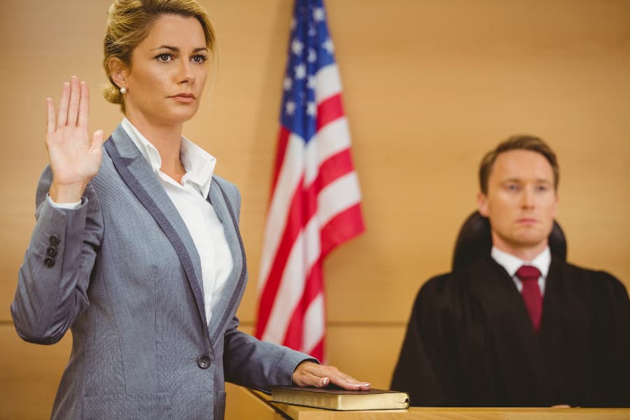 A blonde woman in a suit being sworn in with a judge behind her in a courtroom